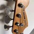 Detail hlavy basové kytary z přírodního dřeva s nápisem Fender MUSTANG BASS. Na hlavě jsou čtyři kovové ladičky s viditelnými známkami opotřebení a o…