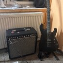Černý kytarový zesilovač značky Fender s nápisem Champion 20. Vedle něj stojí černá elektrická kytara v držáku, na hlavě které je viditelné logo SLT.…