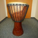 Dřevěný buben typu djembe s hnědým povrchem a světlou koženou membránou. Tělo nástroje je v horní části opletené černým šňůrem a v dolní části má vyř…