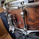 Detailní záběr na snare drum s dřevěným korpusem v hnědé barvě s výraznou strukturou zrnitosti. Na chromovaném obruči je vyryto logo a nápis MAPEX. N…