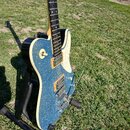 Elektrická kytara s modrým třpytivým povrchem těla a bílým lemováním. Nástroj má dva humbuckery, jeden ovládací knoflík a přepínač. Kytara je umístěn…