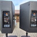 Dvě černé aktivní reproduktory umístěné na stojanech. Na zadním panelu obou kusů je vidět modré logo JBL a nápis PRX800 SERIES. Jsou zde různé vstupy…