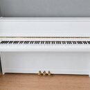 Bílé digitální piano s klaviatura a uzavíracím víkem. Na levé straně nad klávesami je viditelné kulaté logo, které je však nečitelné. Nástroj má zlat…