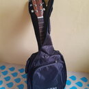 Akustická kytara v černém měkkém pouzdře. Na hlavě kytary je nápis „Sigma Guitars EST. 1970“. Na přední straně pouzdra je bílý nápis „ROCKBAG by Warw…
