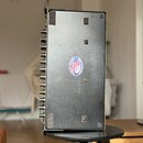 Kovové šasi elektronického zařízení v šedé barvě s viditelnými ovládacími knoflíky na levé straně. Uprostřed spodní strany je kulatá nálepka s americ…