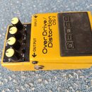 Žlutý kytarový efekt s černým spínacím pedálem. Na horní straně je nápis Over Drive/ Distortion OS-2 a logo BOSS na pedálu. Zařízení má čtyři ovládac…