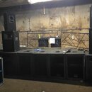 Sestava černých reproduktorů v betonovém prostoru. Jsou vidět subwoofery a topové bedny, z nichž některé nesou logo JBL. V pozadí na stole je umístěn…
