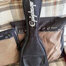 Černý polstrovatý obal na kytaru s bílým nápisem Epiphone®. Obal je umístěn na textilii společně se dvěma černými přepravními sáčky s nápisy Prince A…