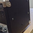 Detail boční strany kytarového komba potaženého černým vinyllem s kovovými rohovými chrániči. Na ovládacím panelu jsou vidět části knoflíků a nápisy…