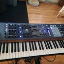 Klávesový synthesizér s tmavě šedým ovládacím panelem a dřevěnými bočními listy. V levém horním rohu je nápis POLYBRUTE a v pravém dolním rohu nápis…