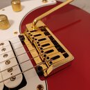 Detail červené elektrické kytary se zlatým kytarovým mostem a pákou. Nástroj má bílý pickguard a bílé snímače. Na zlatém povrchu mostu jsou viditelné…