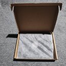 Otevřená hnědá kartonová krabice položená na šedém koberci. Vnitřek krabice je vyplněný bílým ochranným materiálem z bublinkové fólie. Na krabici nej…
