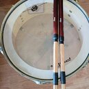 Bílá bubenová blána s nápisem Professional Percussion a logem v čtverci. Na bubnu leží pár dřevěných paliček s červeným lakem v horní části a černými…