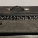 Kytarový zesilovač v černém krytu s kovovou rukojetí. Na ovládacím panelu jsou nápisy INPUT, VOLUME, TREBLE, BASS, GAIN, VOLUME, TREBLE, MID, BASS, R…