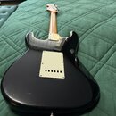 Zadní strana černé elektrické kytary s tvarem typu Stratocaster a dřevěným hřdelem. Na zadní straně těla je světle béžový plastový kryt elektroniky s…