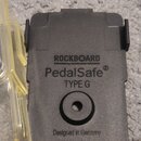 Černý plastový držák s nápisy ROCKBOARD by WARWICK, PedalSafe® a TYPE G. Ve spodní části je text Designed in Germany a kulatý otvor. Povrch plastu je…
