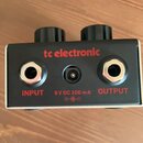 Černý kytarový efekt s červeným nápisem tc electronic. Na zadní straně jsou dva konektory jack a jeden napájecí vstup s nápisy INPUT, OUTPUT a 9 V DC…