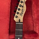 Hlava a hриф elektrické kytary v červeném pouzdře. Na hlavě jsou černé ladičky a nápisy Squier by Fender a TELECASTER. Hриф má tmavý hmatník s bílým…