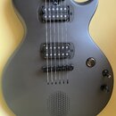 Kytara s matně černým povrchem a dvěma humbuckery. Na těle je vidět mřížka reproduktoru, jeden ovládací knoflík a přepínač. Nástroj nemá žádné vidite…