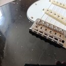 Detail elektrické kytary s černým lakem a bílým pickguardem. Je vidět kovový kobylník s viditelnou korozí a opotřebením povrchu. Na lakovaném těle ky…