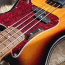Detail elektrické basové kytary s tělem v provedení sunburst. Nástroj má červený členkový pickguard a dva černé snímače. Na těle jsou vidět dvě kovov…