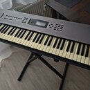 Šedý digitální klavír značky KORG, model N264. Instrument je umístěn na černém X-stojanu a má klasickou klávesnici s bílými a černými klávesami. Na h…
