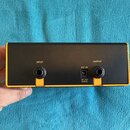Černý obdélníkový box s žlutým okrajem. Na zadní straně jsou dva konektory s nápisy INPUT a OUTPUT oraz středový napájecí vstup s nápisy DC IN a DC 1…