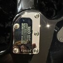 Detail kovového krytu na zadní straně černého nástroje. Na chromovaném povrchu je vyryto logo ERNIE BALL, název MUSIC MAN a nápis CUSTOM MADE. Dole j…