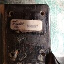 Detail zadní strany černé kytary s viditelným opotřebením laku a nečistotami. Na povrchu je nalepený bílý štítek s logem Fender® a nápisem SERIAL NUM…