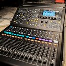 Černý digitální mixážní pult s displejem a řadou posuvníků. V horní části je viditelné logo Midas. Panel obsahuje množství tlačítek, otočných ovladač…