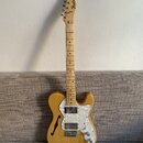 Světle hnědá elektrická kytara s přírodním povrchem dřeva a bílým perleťovým pickguardem. Na hlavě hředu je černý nápis Fender TELECASTER. Tělo kytar…