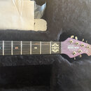 Fialová elektrická kytara v černém plišovém kufru. Na hlavě nástroje jsou bílé nápisy ERNIE BALL, MUSIC MAN a MAJESTY spolu s podpisem. Na hrazdici j…