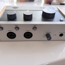 Audio rozhraní v šedém provedení s dřevěnými bočními panely. Na horní straně jsou tři černé otočné ovladače s nápisy GAIN 1, GAIN 2 a MONITOR, v horn…