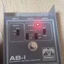 Šedý kovový přístroj s nápisem AB-I BALANCED LINE INPUT SWITCHER a logem Palmer Musical Instruments. Na horní straně jsou označení INPUT A, INPUT B a…