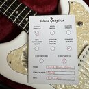 Fotografie zachycuje bílou elektrickou kytaru s perleťovým pickguardem a hnědým hmatníkem. Na kytaru je položen papírový certifikát s nápisem Jolana…
