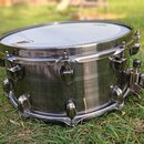 Kovový snare drum s broušeným povrchem a chromovaným kováním. Na bláně je viditelné žluté logo, ale název značky není plně čitelný. Nástroj je v dobr…