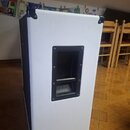 Bílý reproduktor v texturovaném povrchu s černými plastovými rohy. Uprostřed je umístěna černá rukojeť pro přenos. Na přístroji nejsou viditelné žádn…