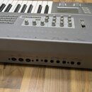 Šedý elektronický klavír s bílými a černými klávesami. Na zadní straně jsou viditelné konektory s nápisy MIDI THRU, MIDI OUT, MIDI IN, DAMPER, ASSIGN…