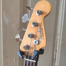 Hlava basové kytary z přírodního dřeva s nápisem Fender PRECISION BASS. Nástroj má čtyři chromované ladičky a je umístěn v černém stojanu. Povrch dře…