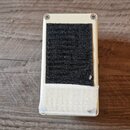 Fotografie zachycuje spodní stranu kytarového efektu v krémové barvě. Na spodku je nalepený černý suchý zip a bílá lepicí páska. Na povrchu nejsou vi…