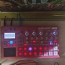 Červený sampler s nápisy electribe sampler a KORG. Zařízení má displej, několik otočných knoflíků a mřížku podsvícených tlačítek. V horní části jsou…