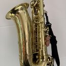 Zlatý saxofon s ENGRAVINGEM na zvonu a v dolní části trubky. Nástroj má černý popruh. Na těle nástroje nejsou viditelné žádné značky ani názvy modelů…