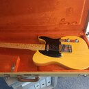 Žlutá elektrická kytara typu Telecaster s černým pickguardem, položená v otevřeném hnědém kufru s oranžovou výplní. Nástroj má světle dřevěný hриф a…
