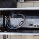 Zadní strana šedého elektronického zařízení umístěného v černém transportním kufru s hliníkovými hranami. Na zadní straně jsou vidět dva konektory SP…