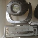 Černý světelný efekt typu moving head s kovovou konstrukcí a čočkou v přední části. Pod ním je šedý ovládací panel s konektory, přepínačem a úchytkou…