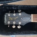 Černá hlava elektrické kytary v černém plišovém kufru. Na hlavě jsou bílé nápisy Epiphone, MODEL Les Paul a Classic. Viditelné je šest ladicích mecha…