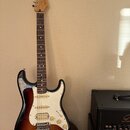 Elektrická kytara s tłem v barvě sunburst, bílým pickguardem a hnědým hmatníkem. Na hlavě je viditelné logo Fender. Kytara má tři snímače, pět úhrado…