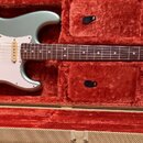 Elektrická kytara s kovově šedým lakem a bílým pickguardem v červeném plišovém kufru. Na hlavě hředu je viditelné logo Fender a nápis STRATOCASTER. K…