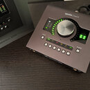 Šedý zvukový interface Apollo Twin značky Universal Audio. Zařízení má velký centrální ovládací knoflík, LED indikátory úrovně a řadu tlačítek s nápi…