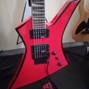 Červená elektrická kytara s ostře hranatým tvarem těla a černými hranami. Nástroj má dva černé humbuckery, jeden ovládací knoflík a přepínač. Kytara…